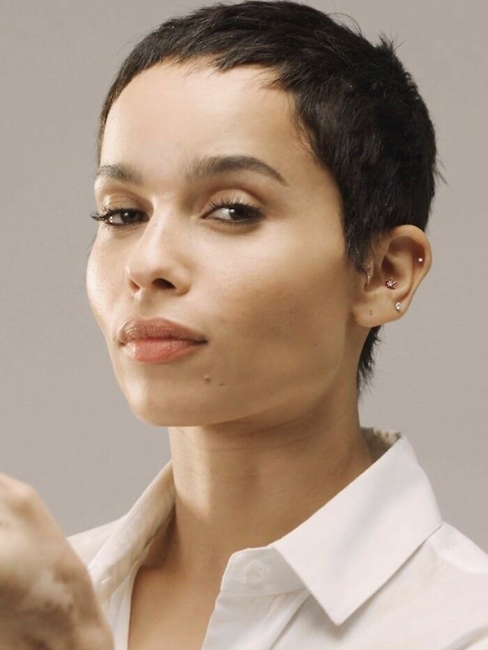 Zoë Kravitz with short hair and multiple ear piercings, wearing a white shirt in a studio portrait.