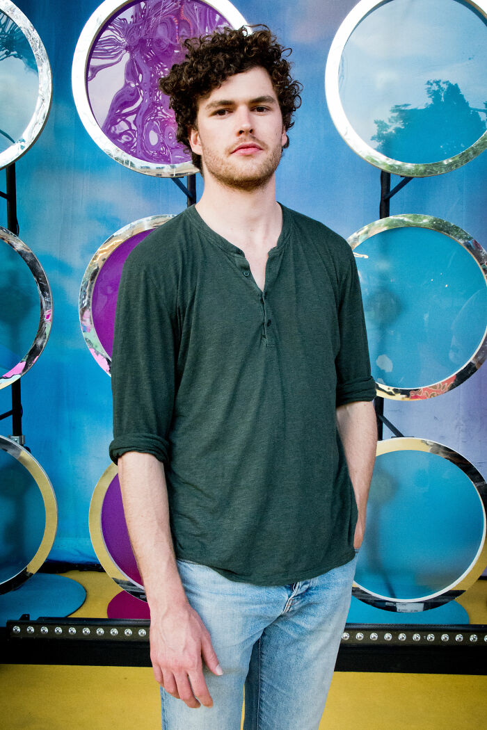 Vance Joy standing in casual outfit with curly hair against a colorful background in a relaxed pose.