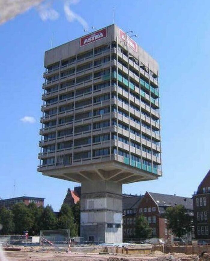 Unusual bizarre building elevated on a narrow base, featuring a boxy design with multiple windows against a clear blue sky.