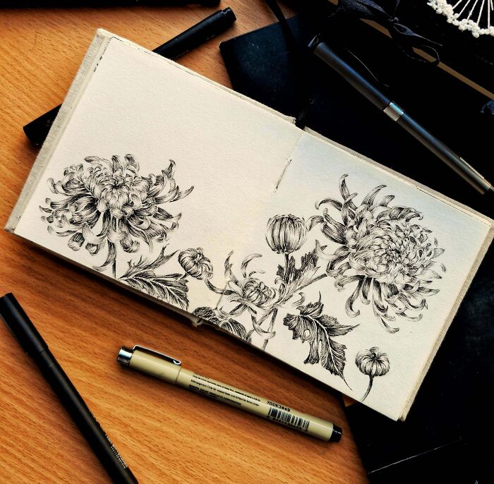 Intricate black ink floral drawing in an open sketchbook with pens on a wooden desk, showcasing cool creative artwork.