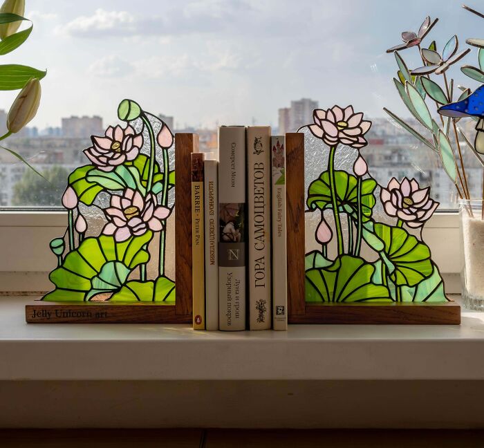Stained glass lotus flower bookends holding books on a windowsill with city buildings in the background, cool handmade art.