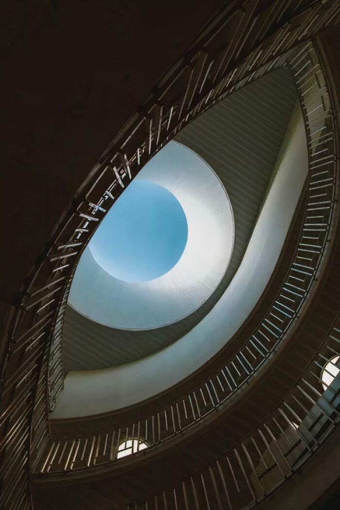 Spiral staircase in an eerie building with curved railings and light shining through a circular opening above.