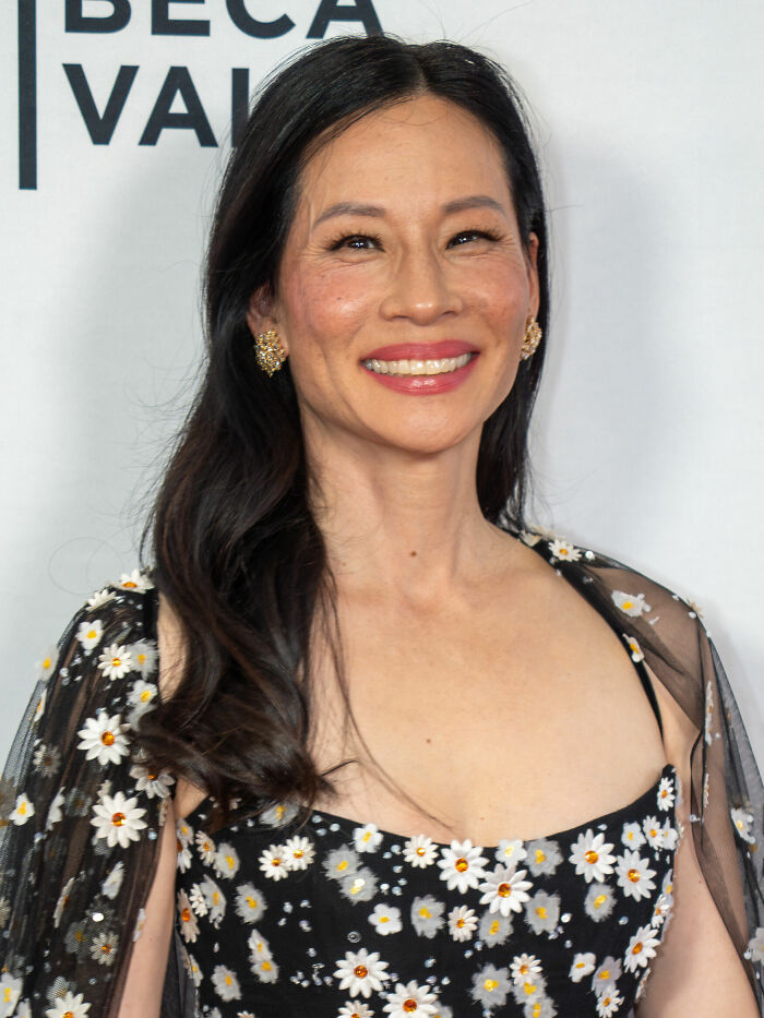 Lucy Liu smiling at an event, wearing a black floral dress with daisies and gold earrings, with long wavy hair.