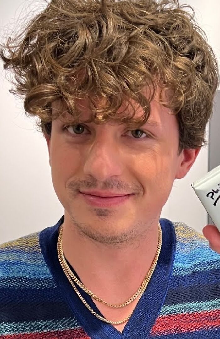 Charlie Puth with curly hair wearing a striped sweater and gold chains, showing a product in a bright indoor setting