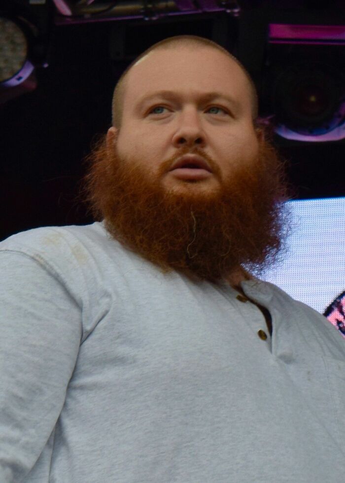 Action Bronson with a full red beard and gray shirt performing on stage during a live music event.