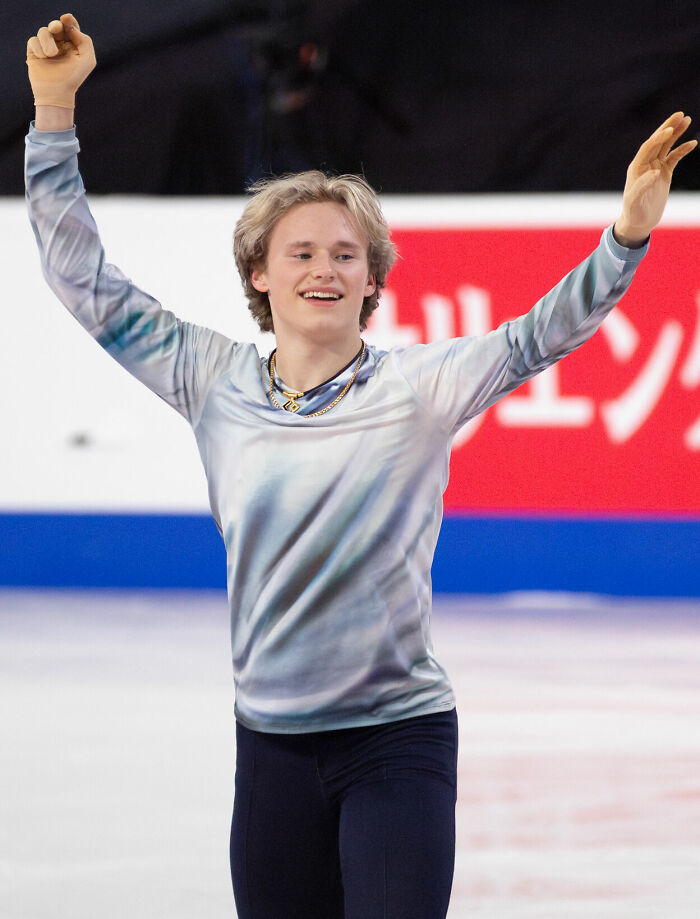 Ilia Malinin smiling and raising arms on the ice during a competitive figure skating performance.