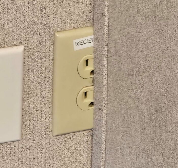Electrical outlet mostly hidden behind a wall panel demonstrating a poorly done job in a workspace setting.