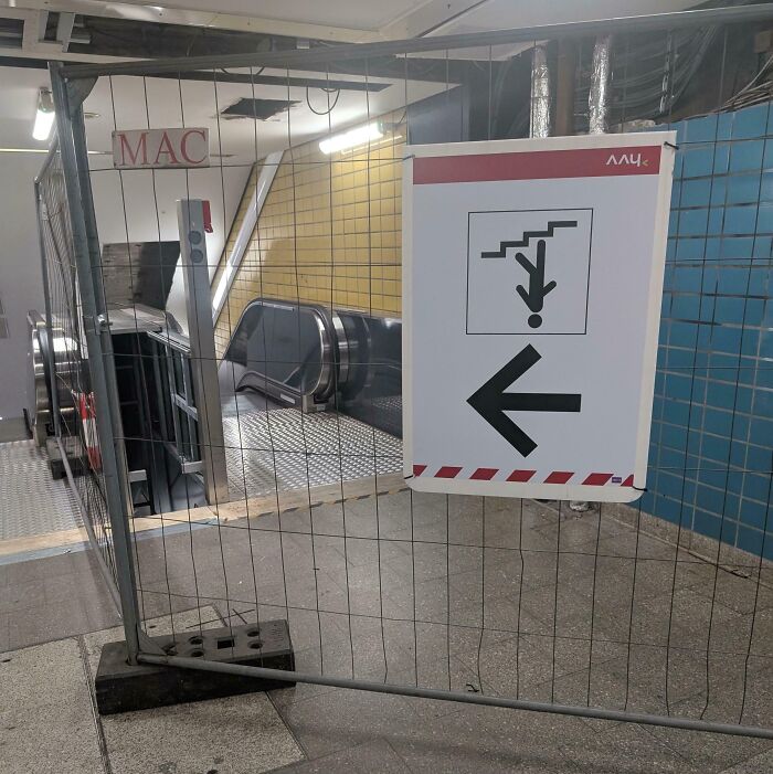 Construction barrier blocking access to escalator and a sign with upside-down figure indicating poor job e*******n.