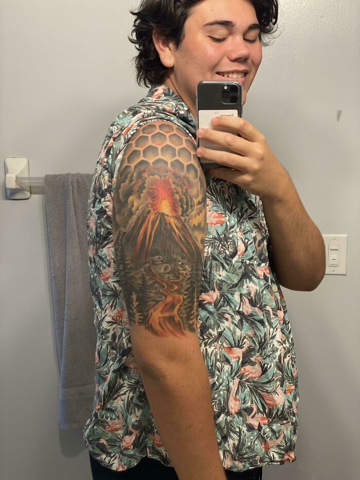 Young man taking a selfie showing a colorful sleeve tattoo with volcano and honeycomb pattern, a wholesome tattoo with deep meaning.