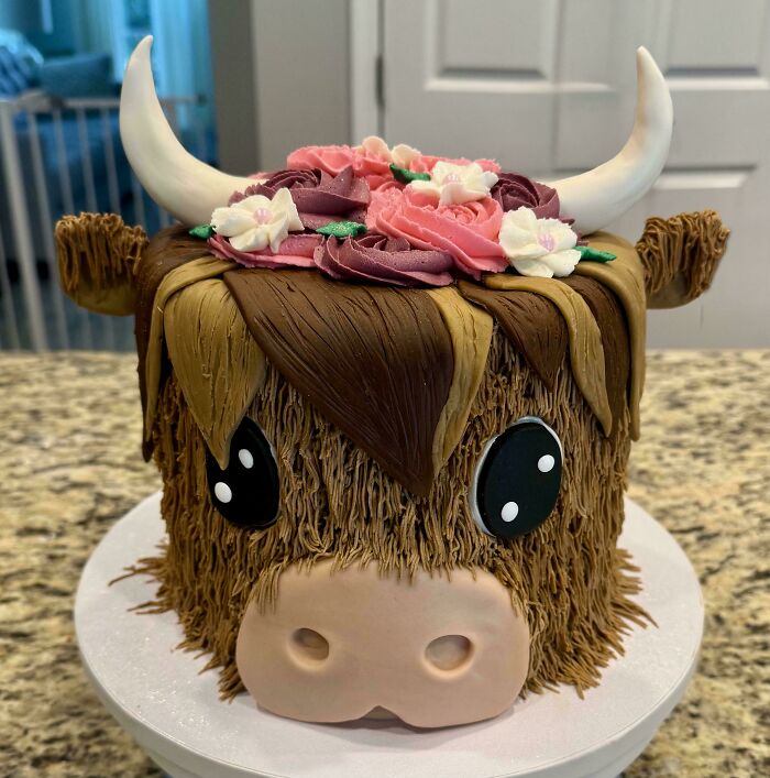 Decorative cake shaped like a highland cow with textured fur and pink flower decorations, showcasing masterfully crafted cakes.
