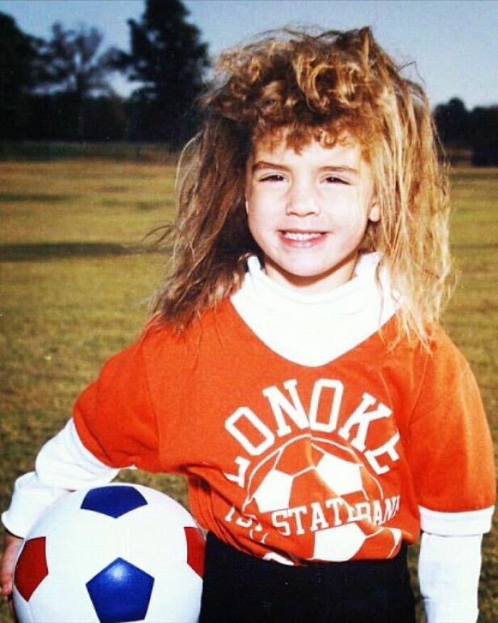 Child with wild hair holding a soccer ball outdoors, capturing a hilariously awkward childhood photo moment.