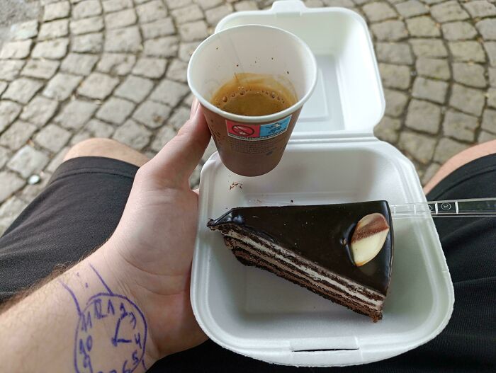Hand holding coffee and cake container with a drawn clock on wrist in a photo so confusing your brain might need a moment to catch up
