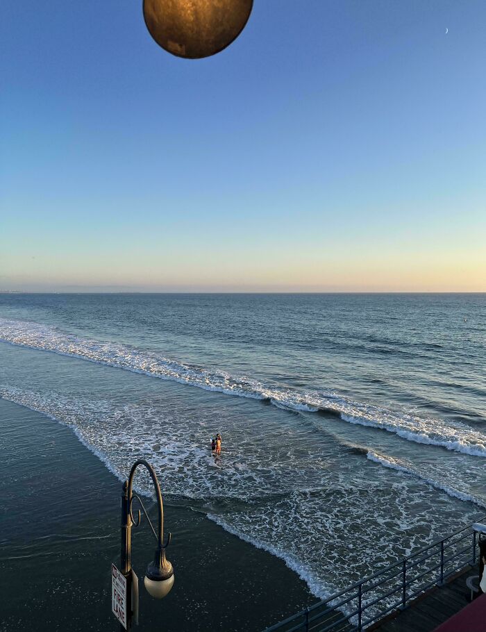 Confusing photo with a large object appearing to hang in the sky over the ocean near a pier at sunset.