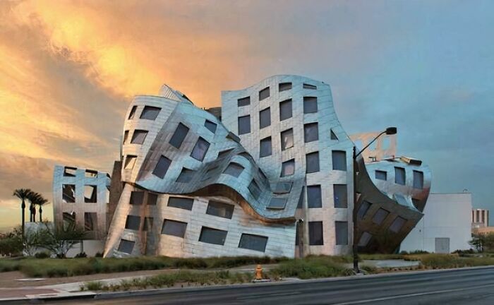 Bizarre building with twisted metal exterior and distorted windows under a colorful sunset sky along an urban street.