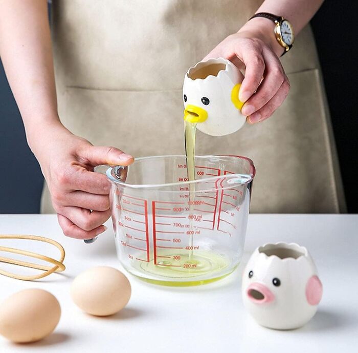 Person cracking egg using a quirky egg separator into a measuring cup, shown as a fun white elephant gift idea.