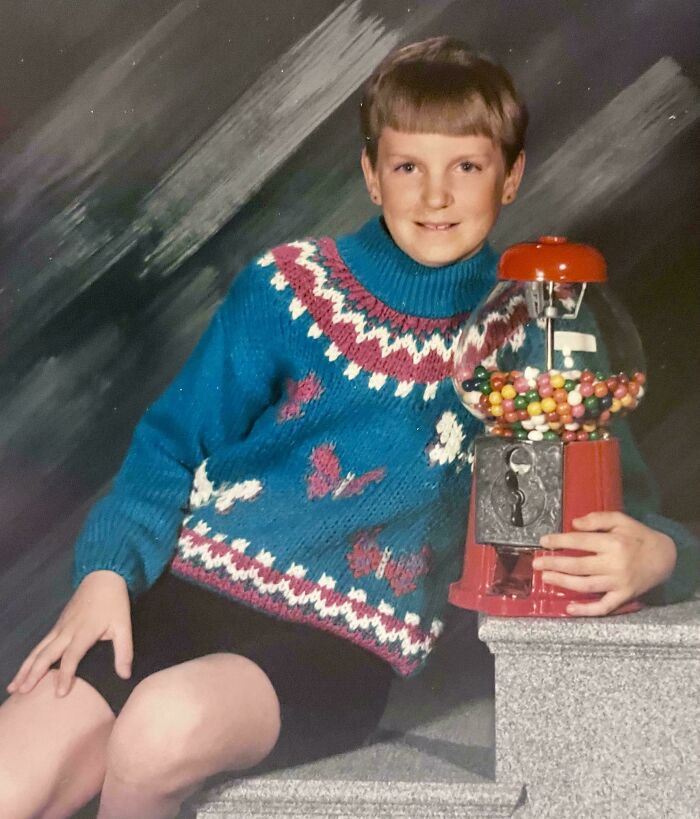 Child in a colorful sweater posing awkwardly with a vintage gumball machine in a retro childhood photo.