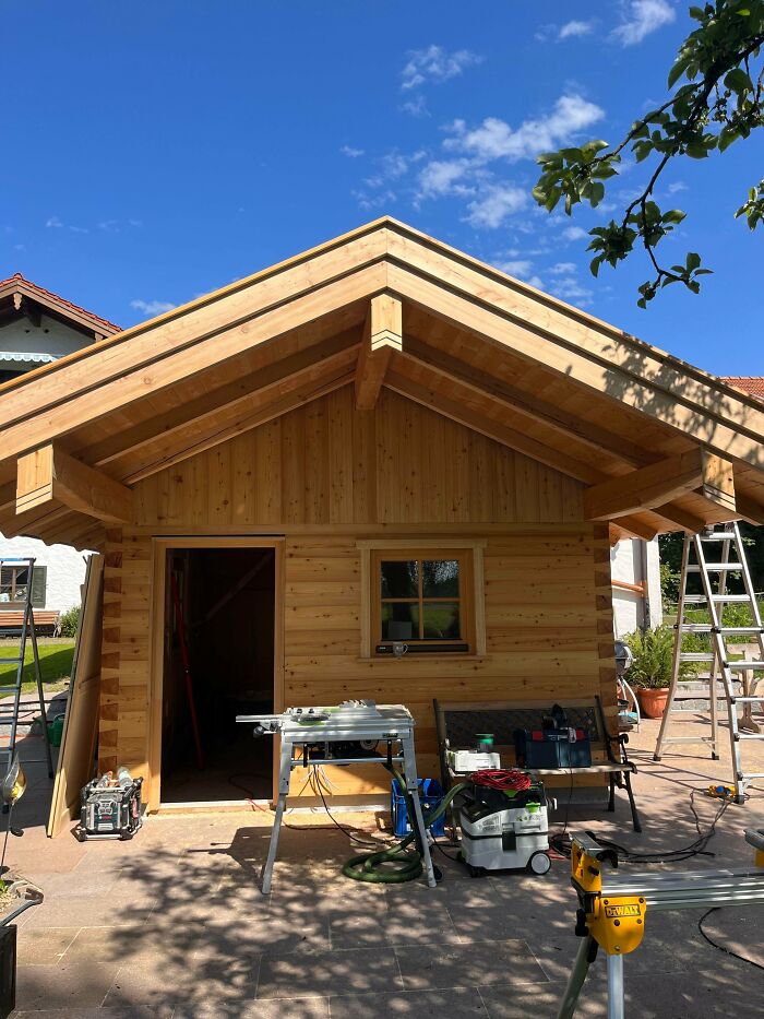 Woodworkers carving a wooden cabin with various tools and equipment under a clear blue sky on a sunny day.