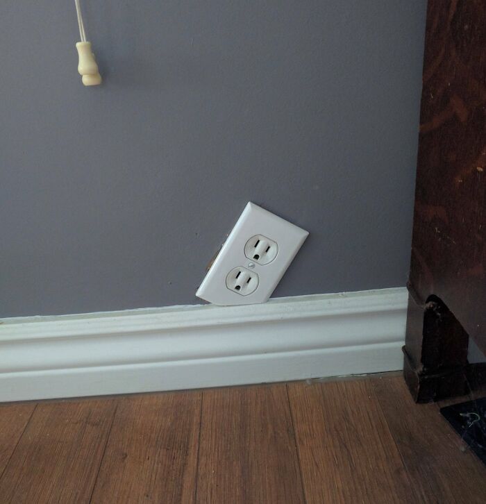 Electrical outlet installed crookedly near the baseboard on a gray wall, showcasing poor workmanship.
