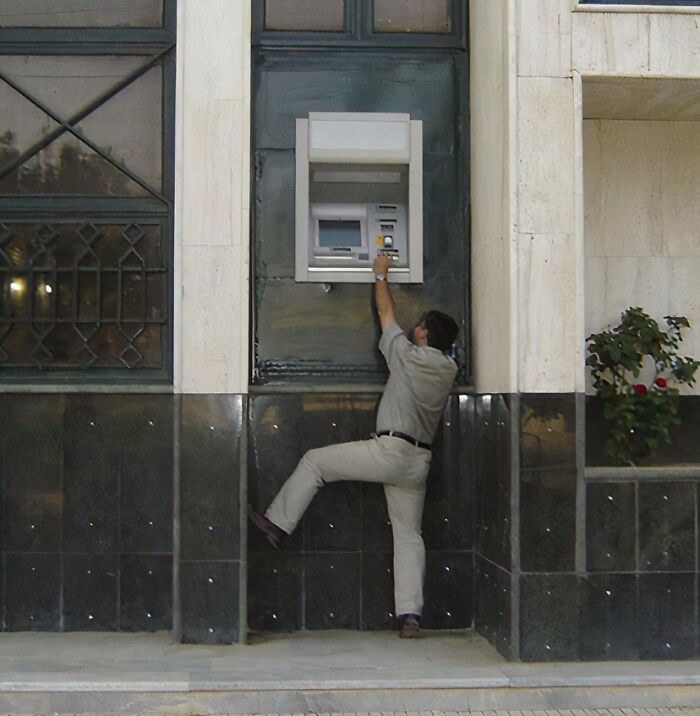 Man struggling to reach an awkwardly placed ATM machine, showing poor job e*******n almost impressive in its failure.