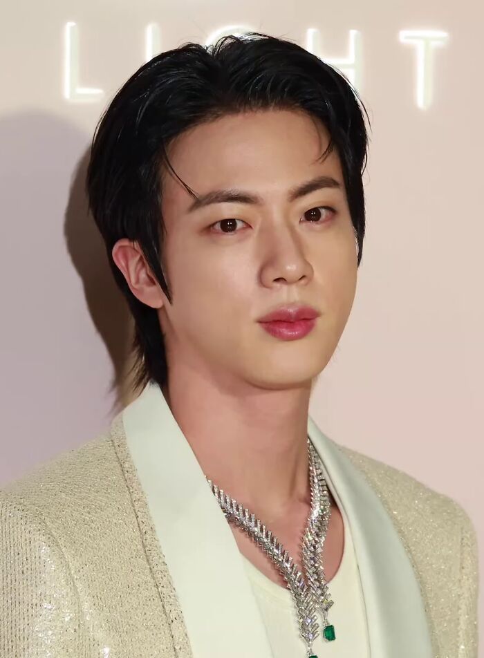 Jin posing at an event, wearing a light-colored jacket and a distinctive silver necklace, showcasing his career highlights.
