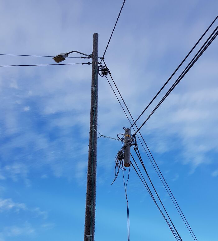Utility pole with tangled and poorly managed electrical wires against a blue sky, showing a job done so poorly it’s almost impressive.