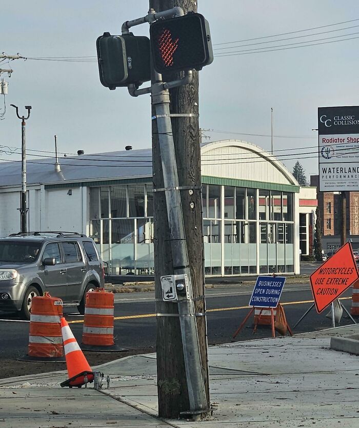 Leaning traffic light pole at a street corner with construction cones and caution signs, showing poor job e*******n.