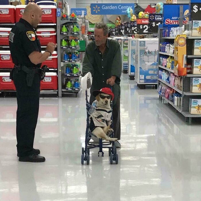 Dog dressed in clothes and sunglasses sitting in stroller while man talks to police officer inside Walmart store aisle.