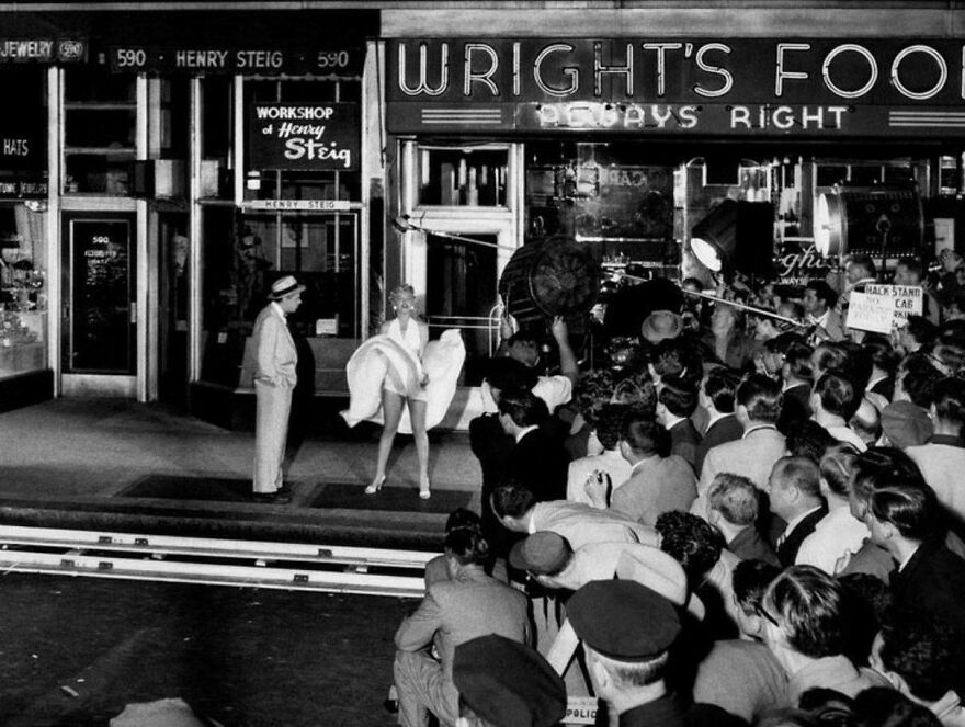 Classic black and white photo of a woman in a white dress attracting a crowd outside a store, an intriguing random picture.