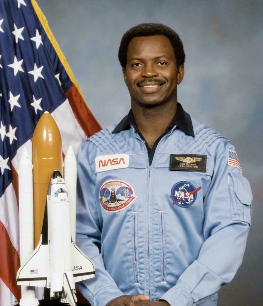 Portrait of a smiling astronaut in NASA uniform standing next to a model space shuttle and American flag, random pictures.