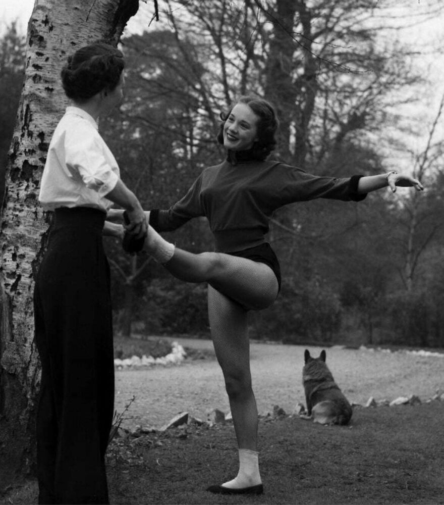 Two women practicing ballet outdoors, one stretching leg with assistance, set in a natural park scene with a dog nearby.