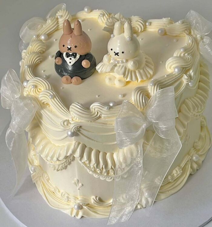 Elegant cream-colored cake with intricate piping, pearl details, and bunny figurines showcasing masterfully crafted cake creativity.