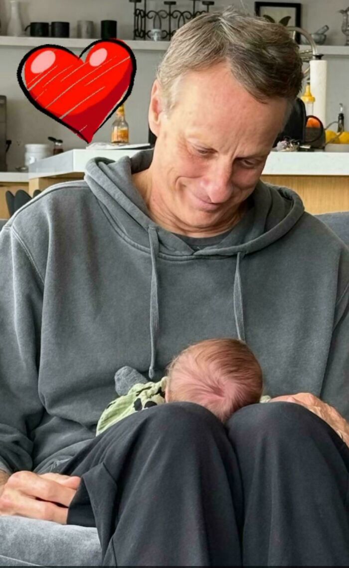 Man in a gray hoodie holding a newborn baby on his lap with a red heart graphic symbolizing heartwarming moments.
