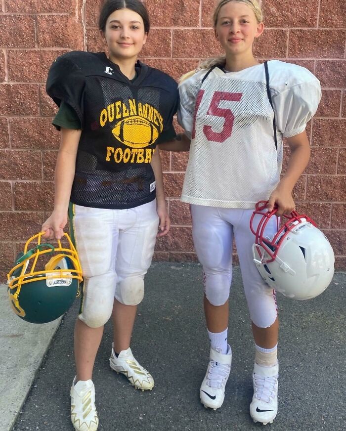 Two young girls in football gear holding helmets, smiling and standing against a brick wall, heartwarming photos.