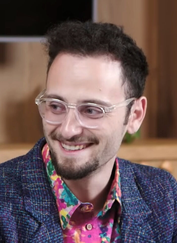 Levy Rozman smiling and wearing clear glasses and a colorful shirt, captured in a casual indoor setting for bio and career highlights.