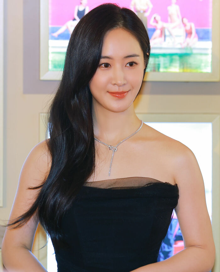 Kwon Yu-ri posing in a black strapless dress with long hair and a silver necklace at an indoor event.