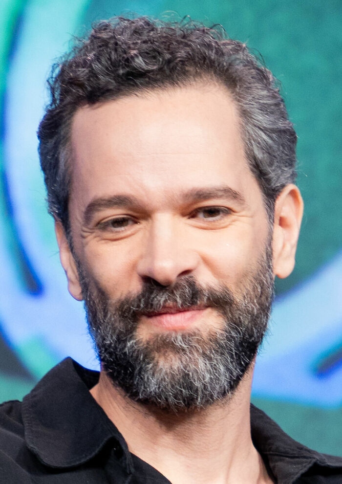 Neil Druckmann with a beard and curly hair, smiling softly against a blurred colorful background.