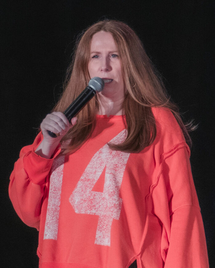 Catherine Tate holding a microphone on stage wearing a bright orange sweatshirt during a live performance.