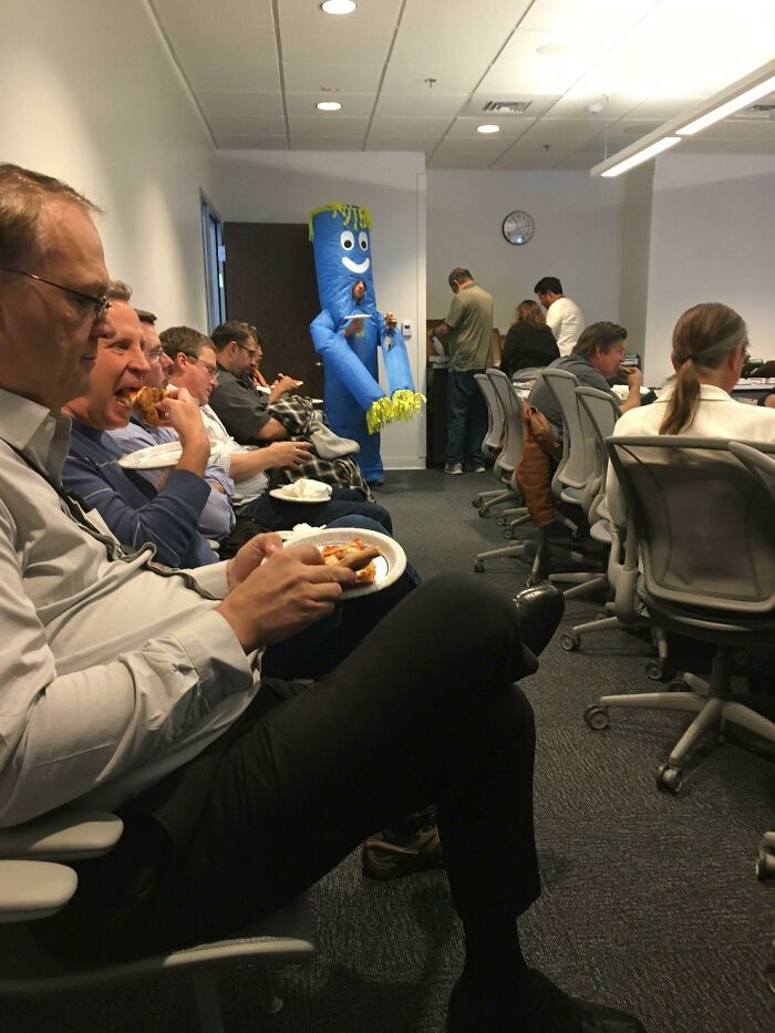 Office workers eating pizza in a meeting room with a person dressed in a blue inflatable costume making a 9-to-5 more bearable.