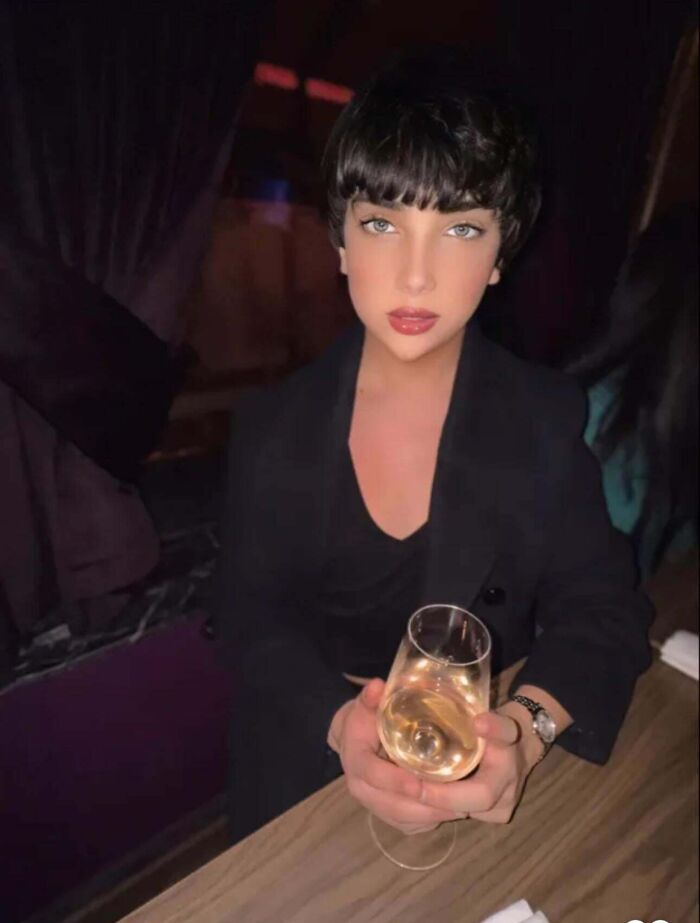 Person holding a glass of wine sitting at a table in dim lighting highlighting Instagram vs reality moments behind glam look.