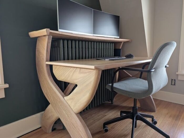 Modern wooden desk carved with unique curved supports, paired with dual monitors and an ergonomic office chair in a home workspace.