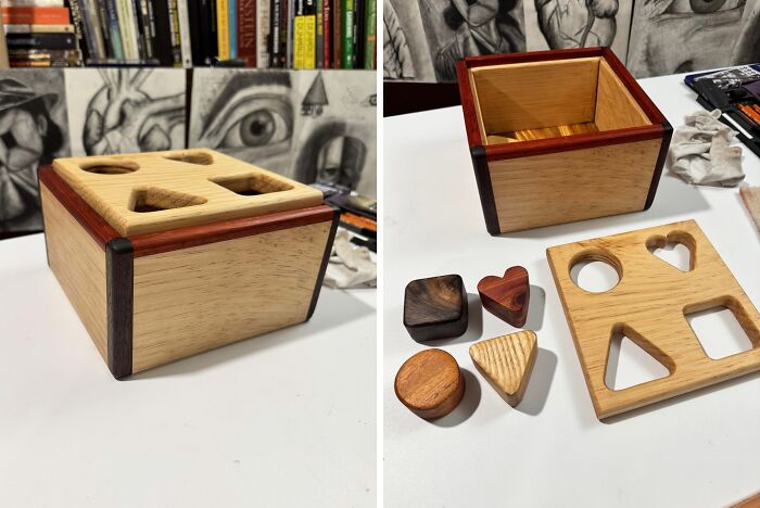Woodworker's handcrafted wooden shape sorter box with various carved shapes on a white surface and books in the background.