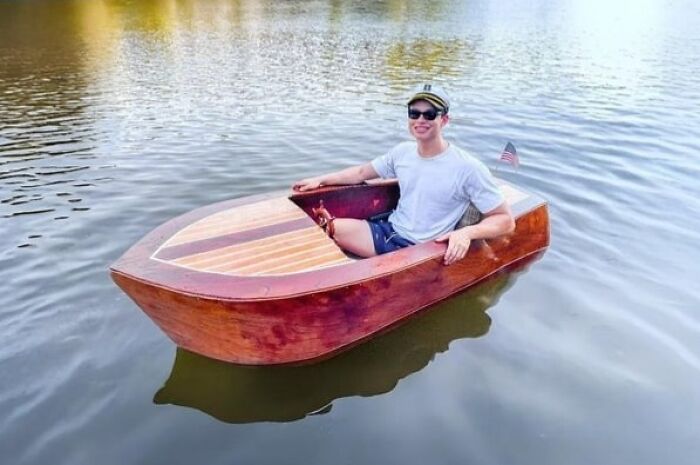 Man wearing sunglasses and a captain's hat sitting in a handmade wooden boat on calm water, showcasing woodworking skills.