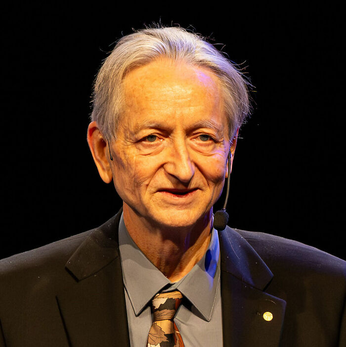 Geoffrey Hinton speaking on stage wearing a suit and tie with a headset microphone in a dark setting.