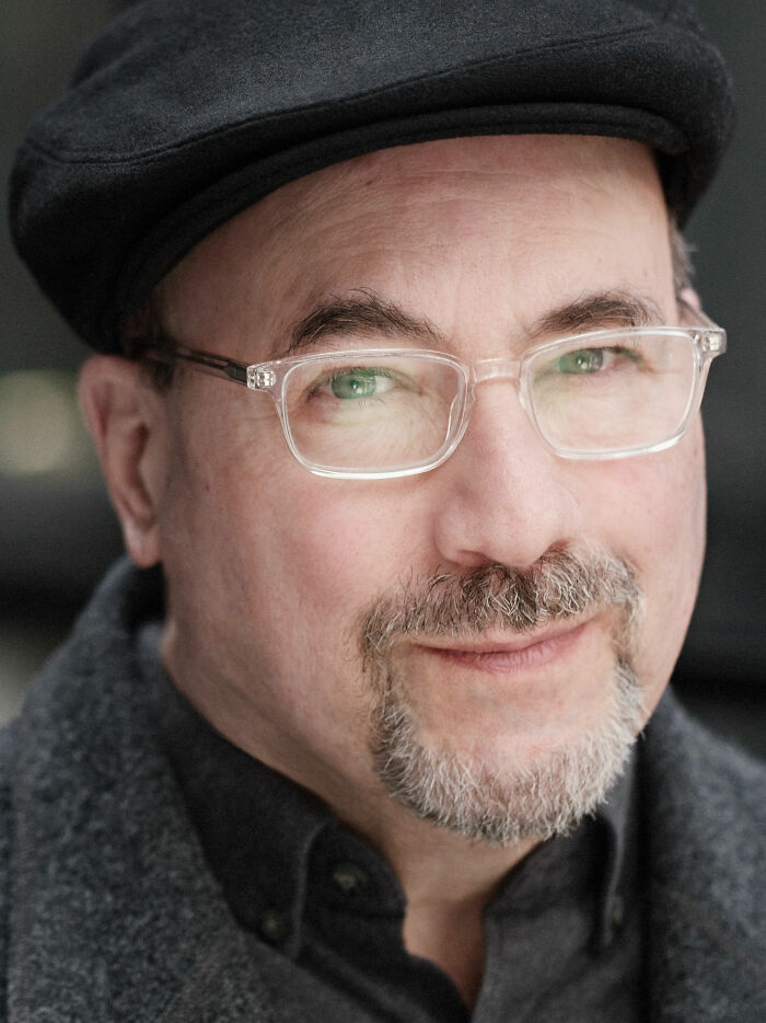 Craig Newmark wearing a black cap and clear glasses with a slight smile, portrait focused on his face.