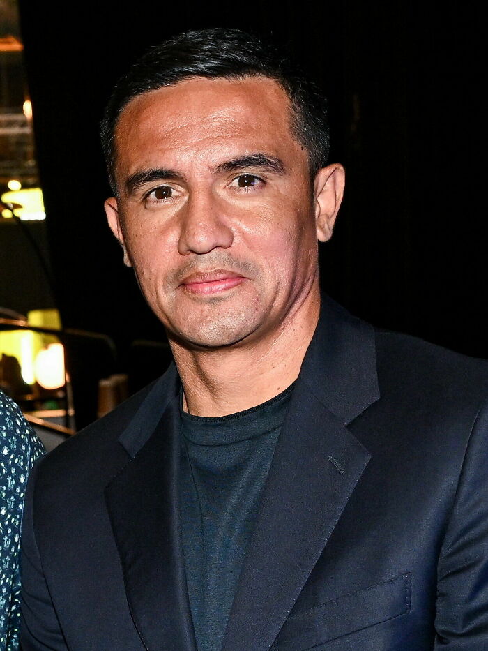 Tim Cahill in a dark blazer and shirt, posing confidently in a dimly lit indoor setting.