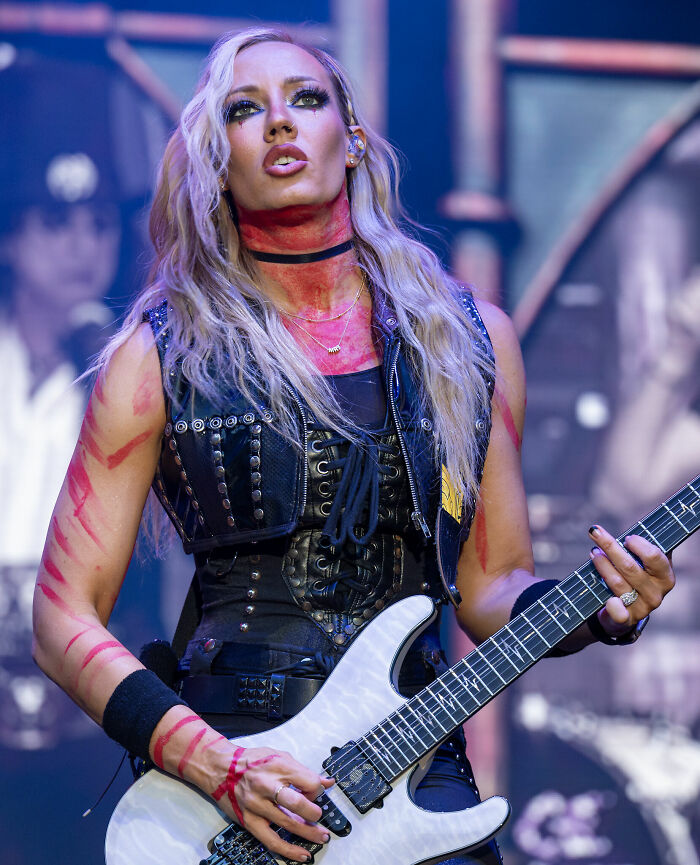 Nita Strauss performing on stage with electric guitar, wearing a studded black outfit and face paint.