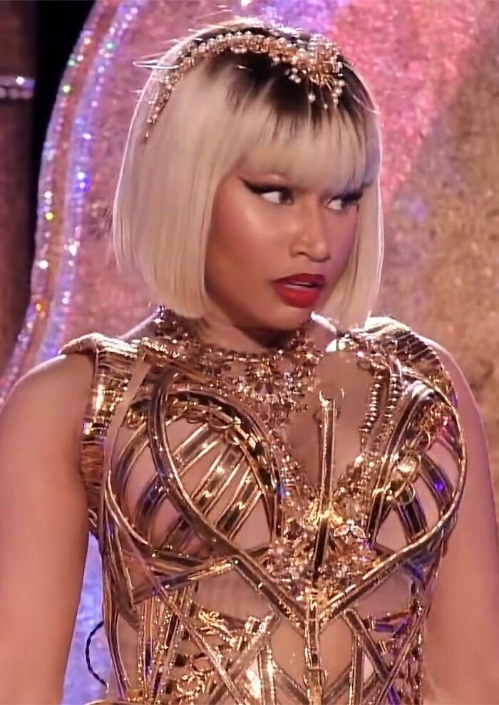 Nicki Minaj wearing a metallic gold outfit with a blonde bob hairstyle and bold makeup during a performance.