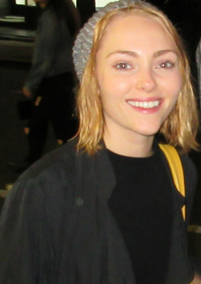 AnnaSophia Robb smiling outdoors, wearing a gray knit hat and dark jacket with a yellow bag strap visible.
