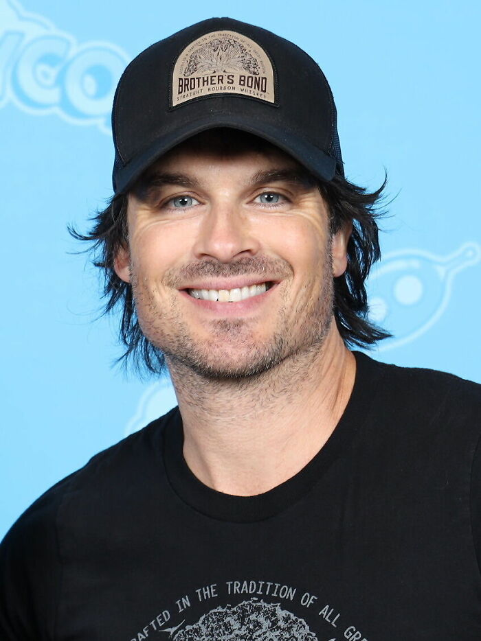 Ian Somerhalder smiling at a fan event wearing a black cap and casual t-shirt with a blue background