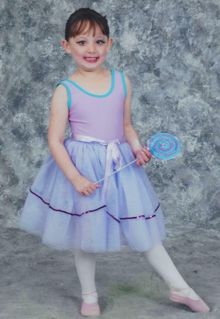 Young girl in a purple tutu dress holding a large lollipop, posing with a shy smile in an awkward childhood photo.